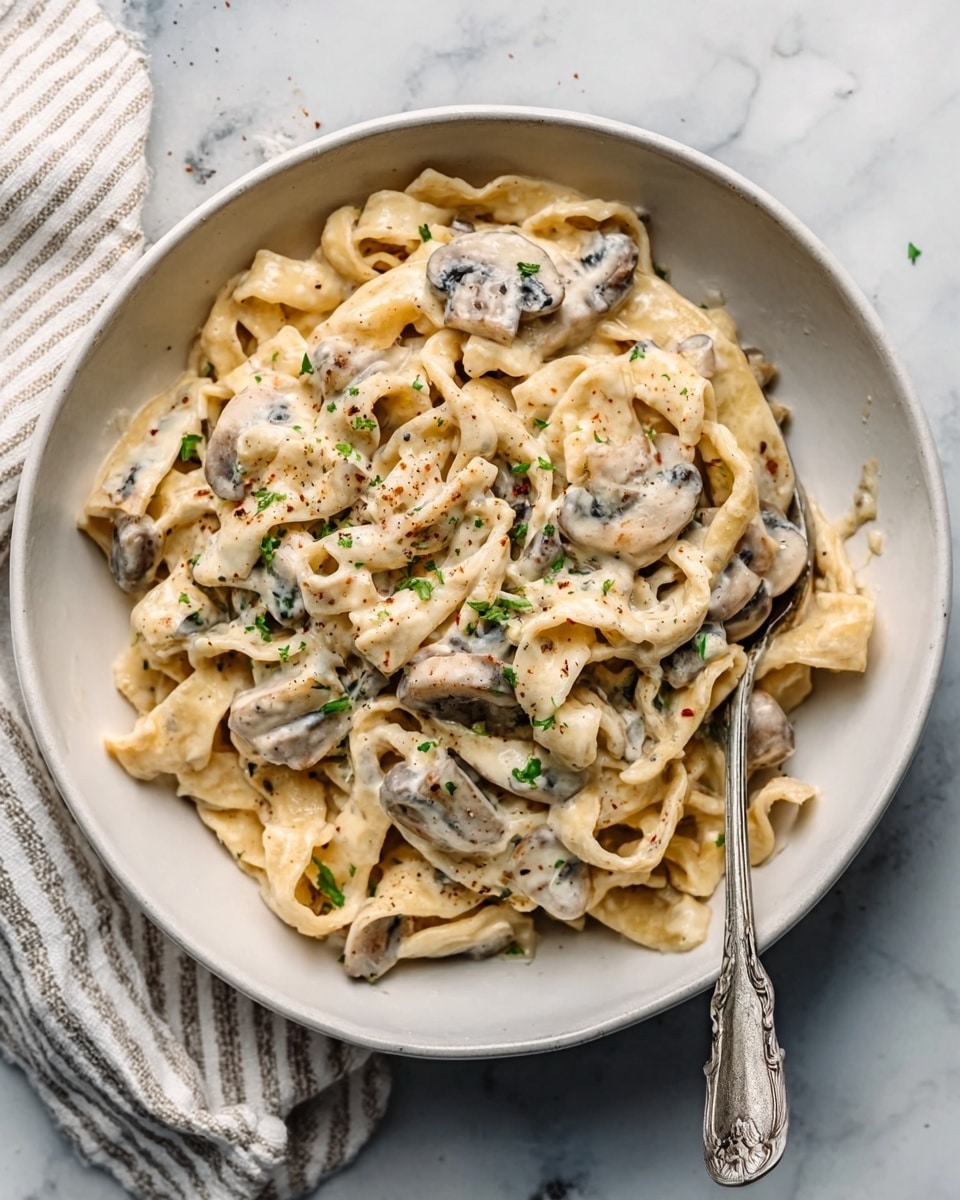 The image shows a white pan filled with cooked penne pasta mixed with pieces of light brown chicken. The pasta and chicken are coated in a creamy white sauce, sprinkled with small green herb pieces for garnish. The pan is placed on a white marbled surface with a red and white checkered cloth nearby. The pasta and chicken are evenly spread in the pan, creating a simple, comforting dish. Photo taken with an iphone --ar 4:5 --v 7