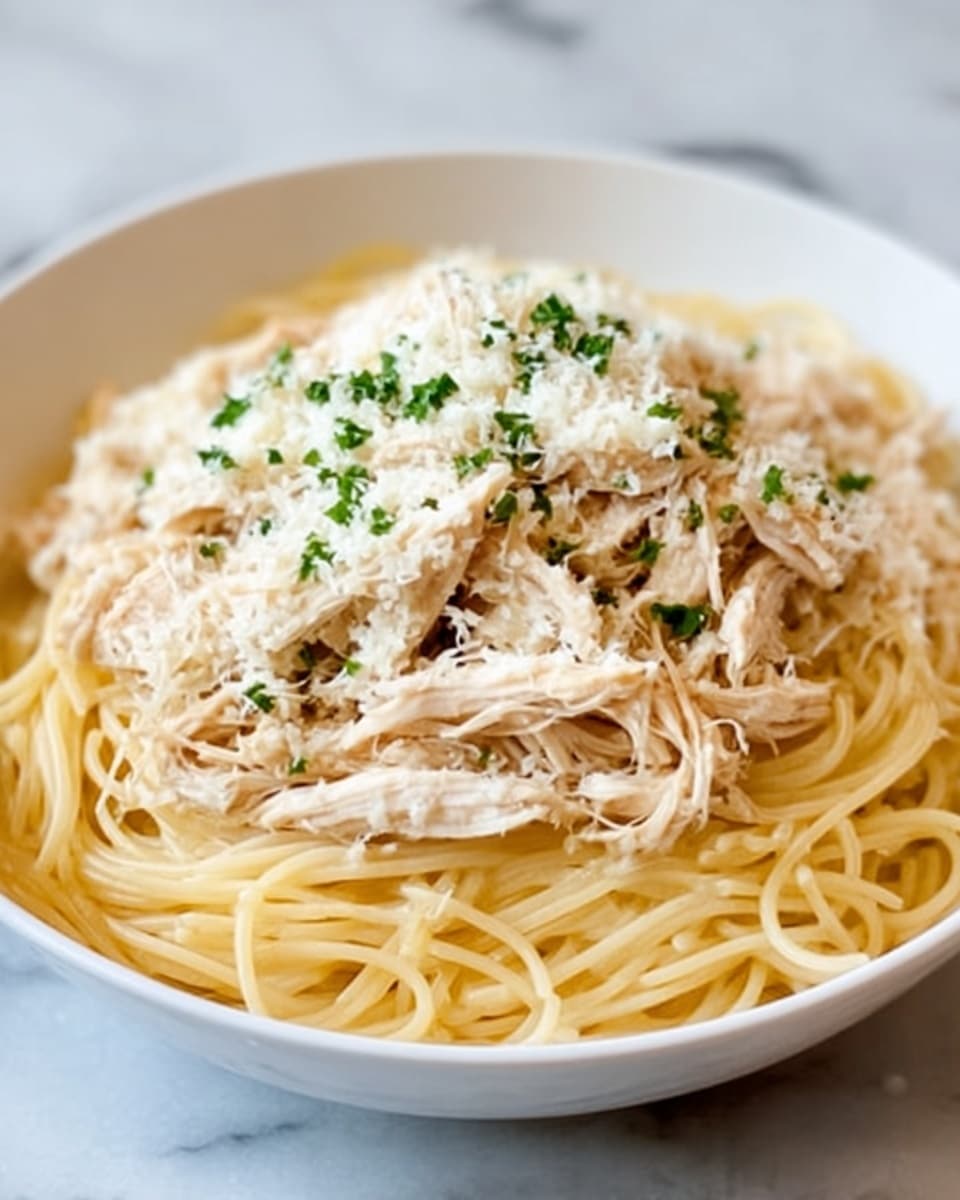 A white bowl holds a dish with a bottom layer of cooked spaghetti, light yellow with a smooth texture, neatly spread to fill the bowl. On top, there is a layer of shredded cooked chicken, pale beige and soft-looking, covering the pasta evenly. The dish is finished with a sprinkling of finely grated white cheese and small green herb bits scattered over the chicken. The bowl sits on a white marbled surface. photo taken with an iphone --ar 4:5 --v 7