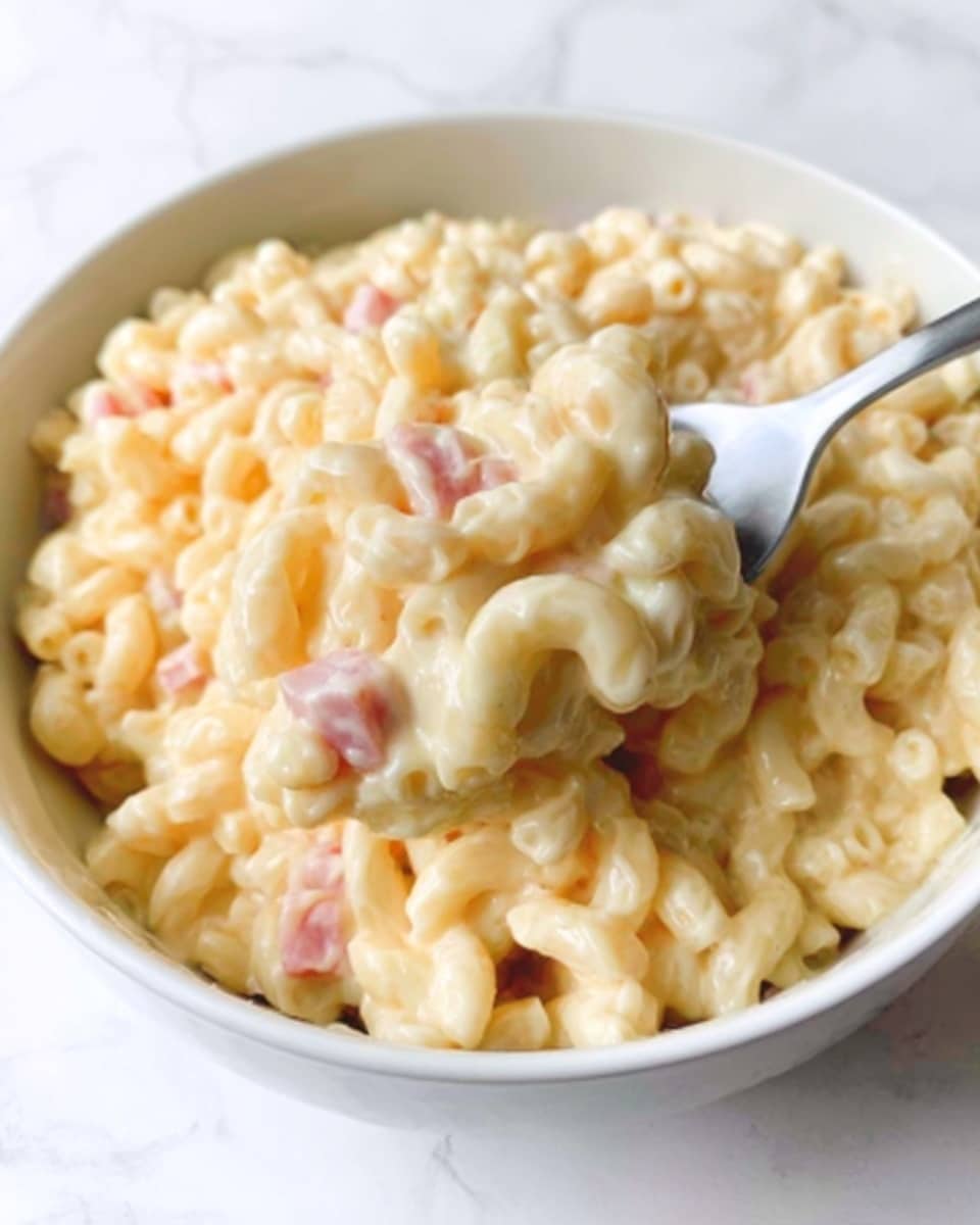 The image shows a close-up of a white bowl filled with creamy macaroni pasta. The macaroni is coated in a smooth, light yellow cheese sauce, with small bits of pink ham mixed throughout. A white spoon is partially visible in the bowl, lifting some macaroni. The bowl sits on a white marbled surface. Photo taken with an iphone --ar 4:5 --v 7