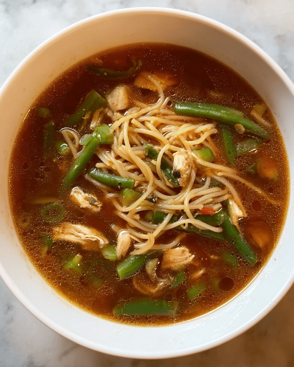 The image shows a white bowl filled with a clear, brown broth soup that has visible oil spots on the surface. Inside the broth, there are thin light beige noodles, irregular small pieces of light brown chicken, and green bean segments scattered throughout. The soup also contains small bits of other vegetables, adding texture and color. The bowl is placed on a white marbled surface. photo taken with an iphone --ar 4:5 --v 7