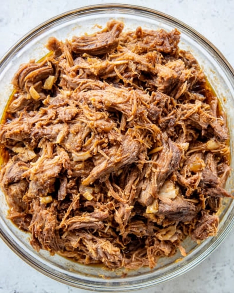 A close-up view of shredded cooked meat placed in a clear glass bowl on a white marbled surface. The meat is brown and looks tender with small pieces of lighter colored bits mixed throughout. The texture appears slightly moist with some visible strands and small chunks layered evenly throughout the bowl. The scene is well-lit showing the rich brown color and varied texture of the meat evenly spread across the bowl. photo taken with an iphone --ar 4:5 --v 7