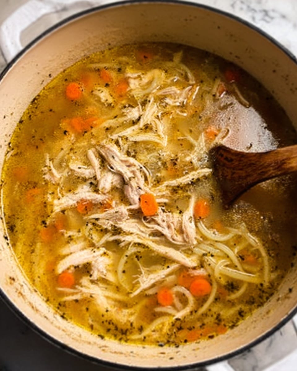 The image shows a large white pot filled with clear, golden broth containing shredded light beige chicken pieces, soft translucent white onion slices, and small orange carrot cubes. The soup is dotted with black specks of herbs and spices, creating texture across the surface. A wooden spoon is partially submerged on the right side, stirring the soup gently. The background features a white marbled texture. photo taken with an iphone --ar 4:5 --v 7