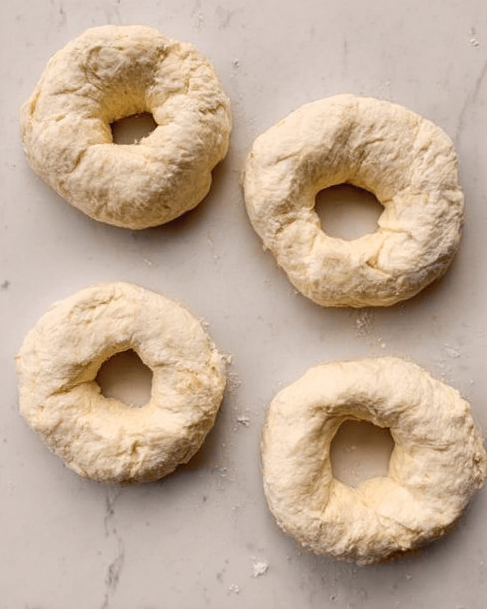 The image shows four raw dough rings placed on a white marbled surface. Each dough ring has a thick, slightly uneven texture with a rough, wrinkled look, and small cracks on the surface. The dough is pale, almost white with a slightly creamy tint. They are spaced apart, with two rings near the top and two near the bottom, forming a loose square shape. The photo is clear and bright, showing the soft, fluffy dough structure. photo taken with an iphone --ar 4:5 --v 7