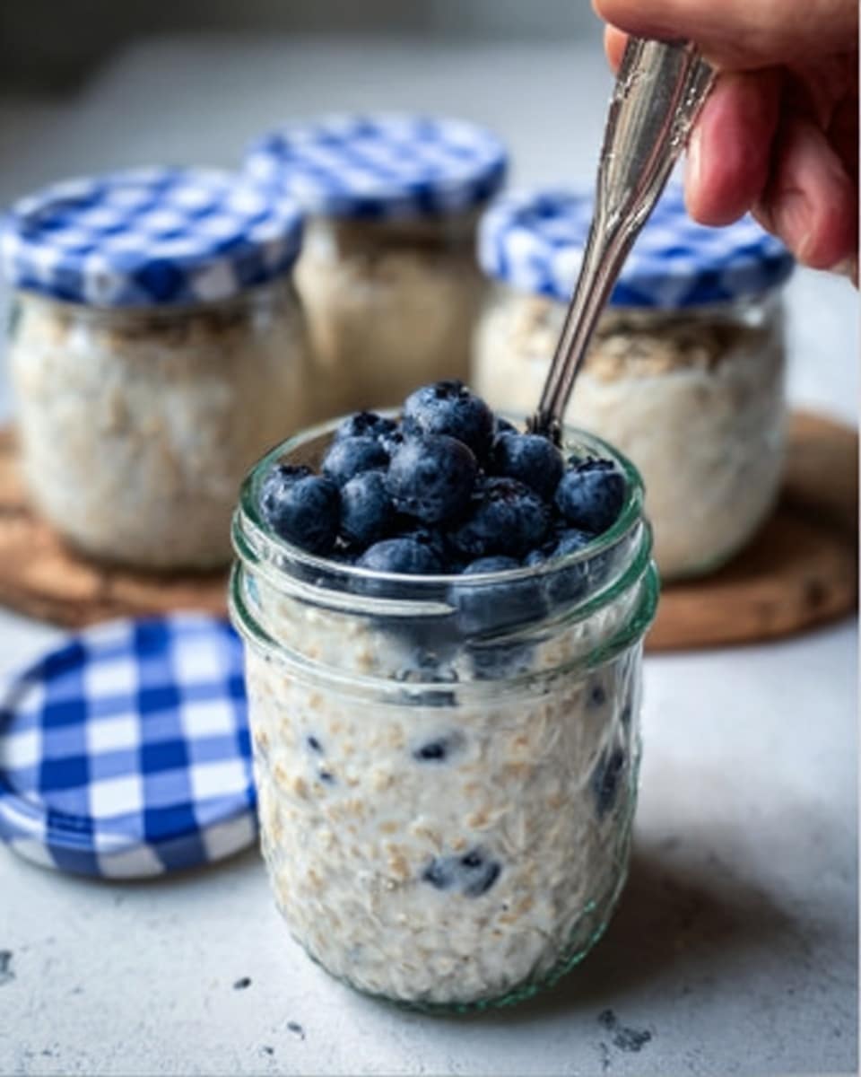 The image shows a clear glass jar filled with creamy oatmeal topped with a layer of fresh, dark blue blueberries spread evenly on top. A silver spoon is inside the jar, partially visible, resting against the side, with a woman's hand holding it. The jar has a blue and white checkered lid placed beside it on a wooden surface. In the background, two similar jars filled with oatmeal and closed with the same lids are slightly out of focus. The overall scene is set on a white marbled textured surface. Photo taken with an iphone --ar 4:5 --v 7