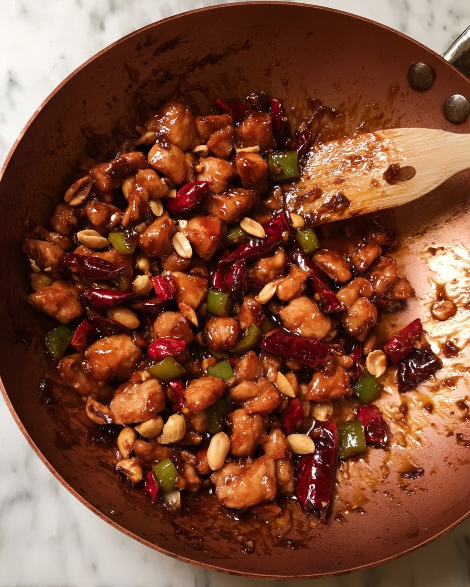 A close-up of a copper-colored pan filled with small, glossy pieces of cooked chicken coated in a dark brown sauce. Mixed throughout are whole peanuts, deep red dry chili peppers, and small chunks of green and red bell peppers, as well as some translucent cooked onions. A wooden spatula rests on the right side of the pan, smeared with the sauce. The pan sits on a white marbled surface. photo taken with an iphone --ar 4:5 --v 7