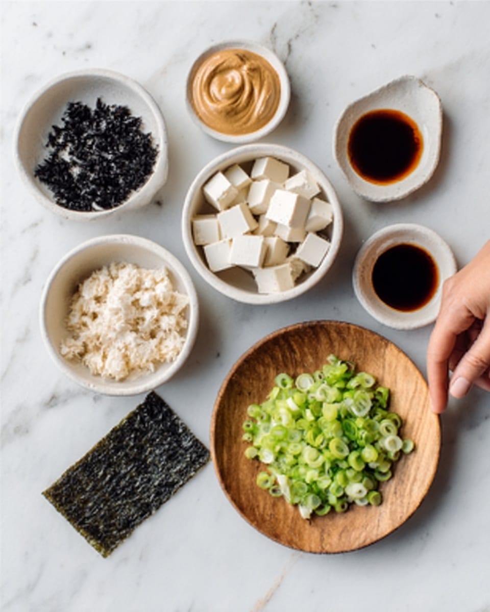 The image shows six small white bowls and a wooden plate placed on a white marbled surface. One white bowl contains white cubed tofu pieces, another white bowl has small black dried seaweed pieces, and a third white bowl holds a light brown creamy paste, likely peanut sauce. The wooden plate is filled with chopped green scallions. A small white bowl nearby holds a dark brown liquid, possibly soy sauce. Next to these, there is a dark, rectangular dried seaweed sheet on the marble surface. A woman's hand reaches into the frame from the right side, lightly touching one of the bowls. photo taken with an iphone --ar 4:5 --v 7