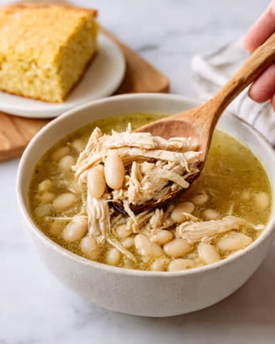 The image shows a white bowl filled with a light yellow broth containing shredded chicken and white beans. A woman's hand is holding a wooden spoon scooping up some of the soup. The broth has visible pieces of tender chicken and soft white beans that fill the bowl. In the background, there is a piece of cornbread on a white plate, all set on a white marbled surface. The colors are soft and warm with a simple, home-cooked feeling. Photo taken with an iphone --ar 4:5 --v 7