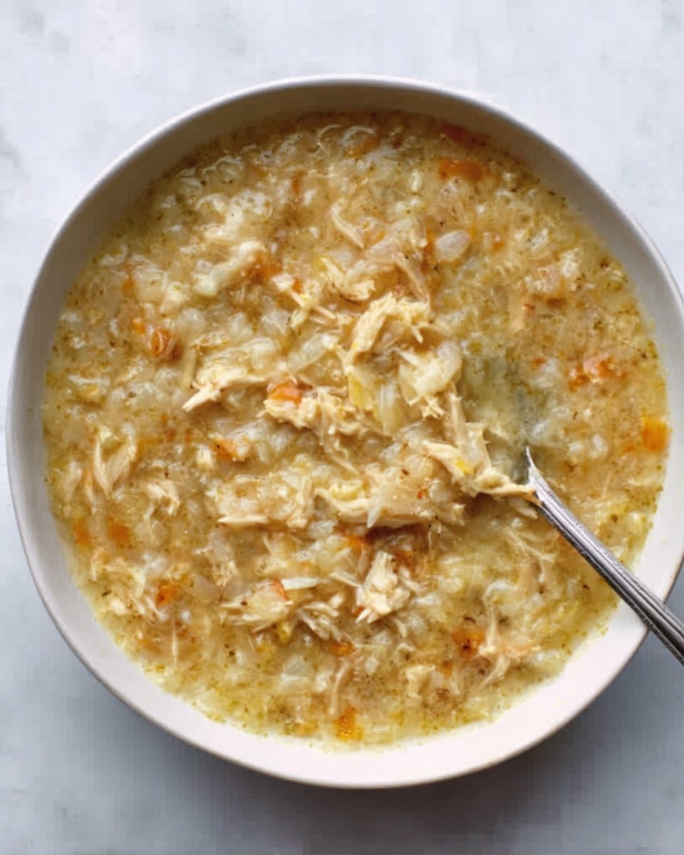 A white round plate with a colorful fruit pattern around the edge holds a creamy beige soup filled with small, soft chunks, likely gnocchi or pieces of dough. The soup is topped with a sprinkle of shredded yellow and white cheese and a light dusting of black pepper. A silver spoon rests inside the plate, partially submerged in the creamy soup. The plate sits on a white marbled surface. The photo taken with an iphone --ar 4:5 --v 7