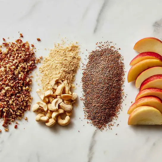 The image shows four neat rows of ingredients on a white marbled surface. On the left, there is a row of crushed pecans with a mix of brown and cream colors, next to it a row of chopped cashews in light cream shade. To the right, there is a row of finely ground flax seeds, dark brown and fine in texture. On the far right, there is a stack of thin apple slices, fresh with red and yellow skin showing on the edges, stacked neatly in a slightly curved line. Photo taken with an iphone --ar 4:5 --v 7