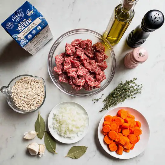 The image shows a clear white marbled surface with several glass bowls arranged neatly. At the center is a large bowl filled with raw red beef pieces. Surrounding it are smaller bowls with bright orange carrot slices, pale green celery pieces, finely chopped white onions, creamy garlic cloves, white grains (likely barley), and vibrant red tomato paste. There are also small bowls with coarse black pepper, a mixture of seasonings, soy sauce, and a cup of dark brown broth. The bowls are all white and clean, and the lighting is soft and natural, highlighting the fresh ingredients clearly. Photo taken with an iphone --ar 4:5 --v 7