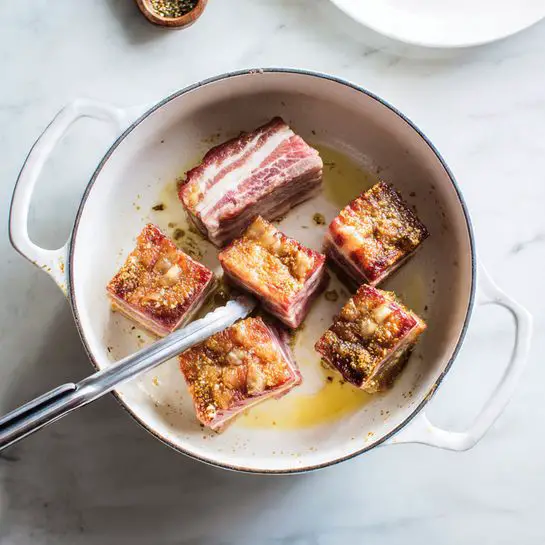 The image shows two white cooking pots on a white marbled surface. In the top pot, there are five raw meat pieces with pink and red tones, some with visible white fat and bone, lying flat in a light oil coating. The bottom pot contains four cooked meat pieces with golden brown crispy skin being turned with silver metal tongs. The pot has browned bits stuck to the sides and bottom from cooking. A white plate is partially seen at the corner near the bottom pot. photo taken with an iphone --ar 4:5 --v 7