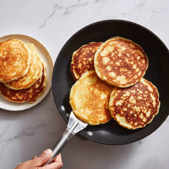 The image shows four golden brown pancakes cooking in a black non-stick pan, with a woman’s hand holding a metal spatula lifting one pancake. The pancakes have a slightly rough texture with small bubbles on the surface. Below the pan is a white plate piled with several cooked pancakes, stacked unevenly with a soft, fluffy look and light brown patches. The background is a white marbled surface. Photo taken with an iphone --ar 4:5 --v 7