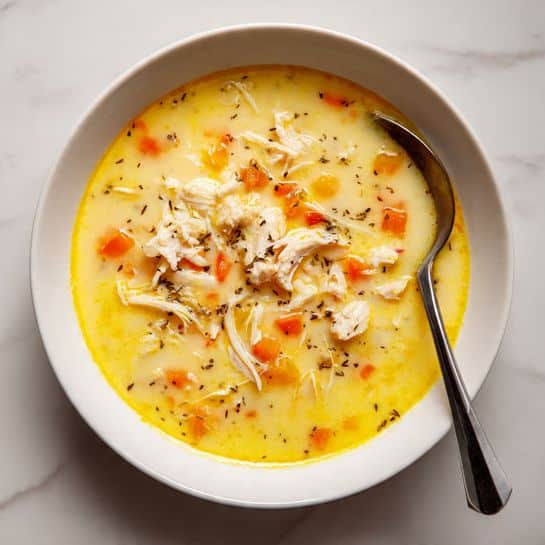 The dish is served in a white bowl placed on a white marbled surface. It has three main layers: the bottom layer is a thick yellow broth with bits of orange carrot pieces scattered throughout, the middle layer consists of shredded white chicken mixed into the soup, and the top layer shows small brown and orange chunks floating on the surface. A spoon is resting on the edge of the bowl, partially dipped into the dish. The texture looks soft and comforting, with the colors blending gently but visibly separated. photo taken with an iphone --ar 4:5 --v 7