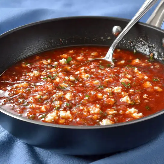 A black pan filled with a chunky red sauce mixed with small white cheese bits on top, showing creamy texture and small green herb pieces scattered throughout. A silver spoon is inside the pan stirring the sauce. The pan is placed on a blue cloth with a soft texture. photo taken with an iphone --ar 4:5 --v 7