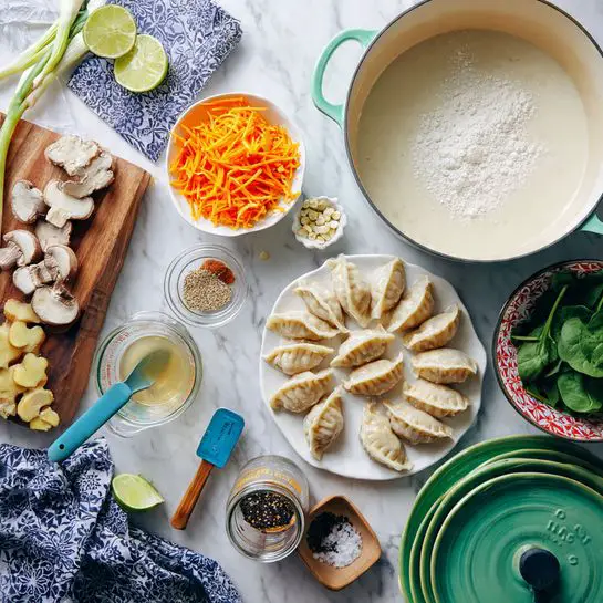 The image shows a cooking setup on a white marbled surface with fresh ingredients and kitchen tools arranged neatly. On the top right, there is an empty light green pot with cream inside. Below it, on the right side, there is a white plate with red trim holding a single layer of half-moon shaped dumplings dusted with flour. Next to the plate, on the bottom right, is a small white bowl full of fresh spinach leaves. In the center, there is a glass measuring cup filled with broth, a small jar with dark chili flakes and a blue measuring spoon, and a small wooden plate with black sesame seeds and a blue measuring spoon. On the left side, a green cutting board holds cut shiitake mushrooms, a halved lime, and three garlic cloves with a knife lying flat. Above the cutting board is a small round white bowl filled with shredded orange carrots, sitting on a stack of blue and green plates with a large piece of fresh ginger resting on top. At the bottom left corner, there are some green onions and a set of light green measuring cups. A blue patterned cloth is folded next to the pot on the marble surface. The composition is bright and colorful with fresh, raw ingredients and cooking tools ready for use photo taken with an iphone --ar 4:5 --v 7