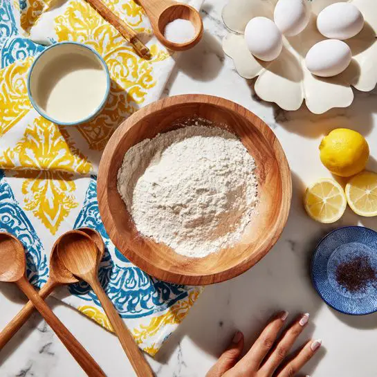 A wooden bowl filled with flour sits on a white marbled surface decorated with a yellow and blue patterned cloth underneath. Three white eggs rest on a white plate shaped like a flower to the right. Nearby are two small lemon halves, a small white bowl with creamy white liquid, and a small blue saucer holding a dark brown ingredient. Three wooden spoons are placed near the bottom left corner. A woman's hand is gently touching the edge of the patterned cloth. The scene is bright and warmly lit. photo taken with an iphone --ar 4:5 --v 7