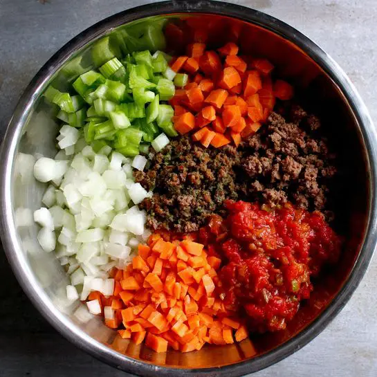 Inside a silver metal bowl, five different food ingredients are placed in separate sections. Starting from the left, there are small chopped green celery pieces, next to them on the bottom left is a pile of white chopped onion, in the center bottom is a mound of finely diced orange carrots, above the carrots to the right is bright red crushed or diced tomatoes, and on top in the middle is dark brown ground meat or similar. Each ingredient is clearly separated and shows its distinct color and texture against the shiny metal bowl. The photo taken with an iphone --ar 4:5 --v 7