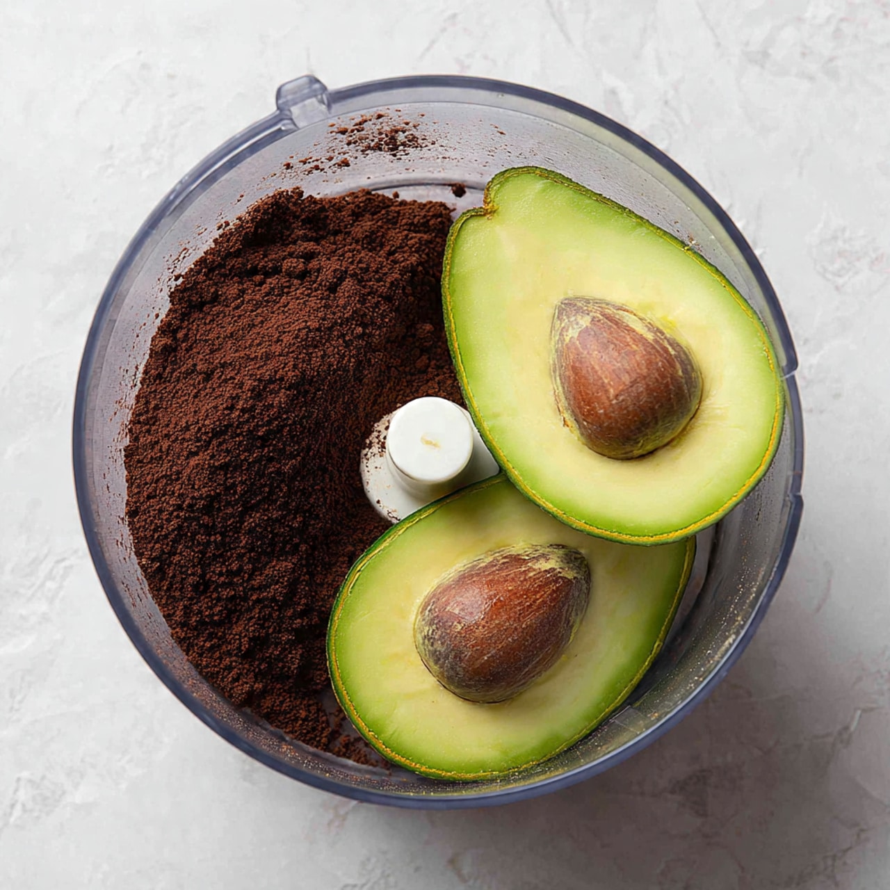 In a clear food processor bowl with white edges, there are two light green avocado halves placed side by side with their brown pits removed, showing the soft avocado flesh inside. On top of one avocado half, there is a heap of dark brown cocoa powder with a powdery texture, covering part of the avocado and resting against a white center blade. The background shows a white marbled surface. Photo taken with an iphone --ar 4:5 --v 7