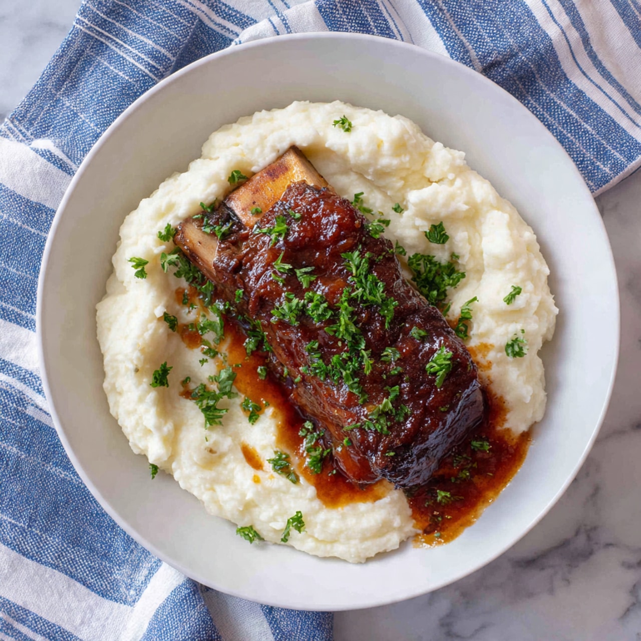 A white plate holds a serving of smooth, creamy white mashed potatoes spread in an irregular oval shape on the bottom left. On top of the mashed potatoes lies a rich, dark brown braised beef rib with a shiny, thick sauce covering it. Small pieces of bright green chopped parsley are sprinkled over the meat and mashed potatoes. A bit of the sauce pools on the plate near the rib. The plate is on a blue and white striped cloth with a white marbled surface underneath. photo taken with an iphone --ar 4:5 --v 7