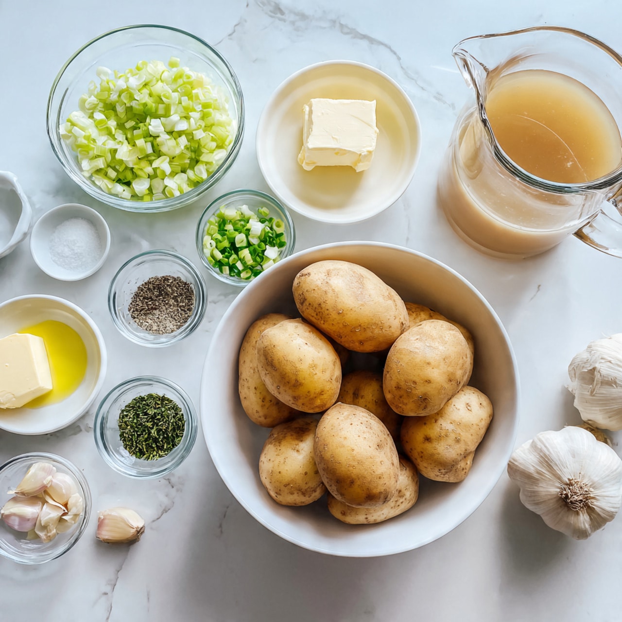 The image shows a white bowl full of several brown potatoes in the center of a white marbled surface. Around the bowl, there are small clear bowls and a glass pitcher, each holding different ingredients: chopped light green leeks in a clear bowl, a small amount of pale yellow butter on a white plate, pale yellow oil in a small clear bowl, two peeled garlic cloves next to a small clear bowl of green herbs, and two small white bowls with black and white pepper. The large glass pitcher contains light brown broth. Photo taken with an iphone --ar 4:5 --v 7