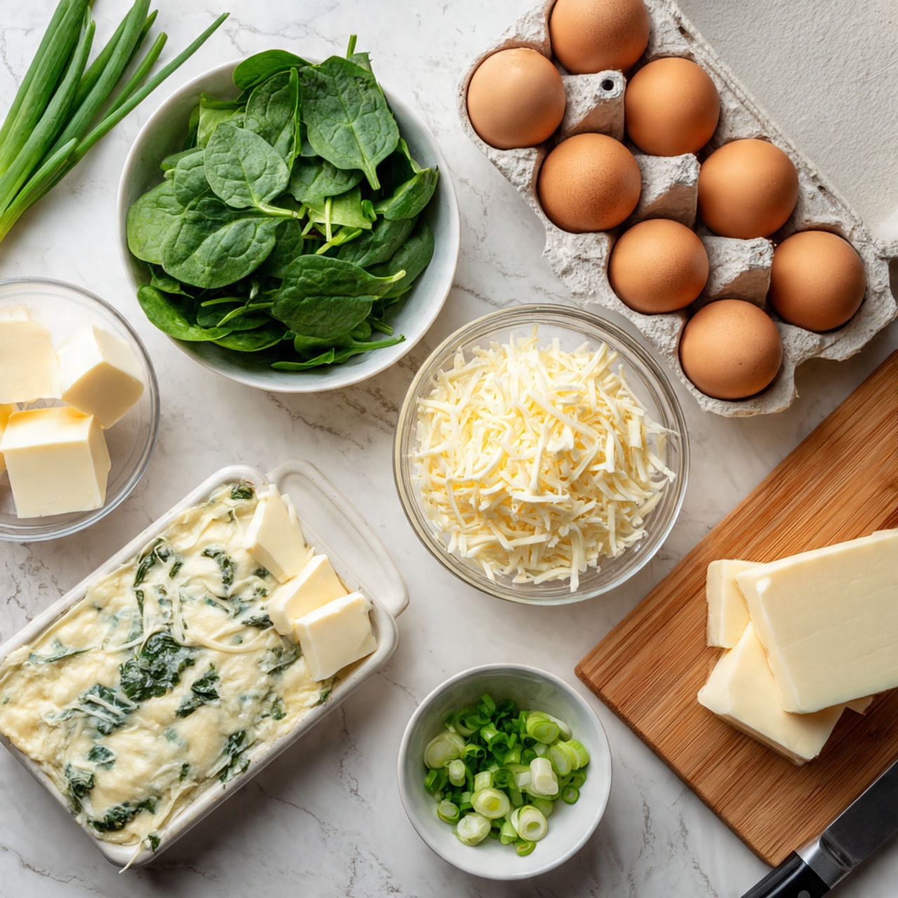 The image shows ingredients on a white marbled surface arranged neatly, including a small white bowl filled with fresh green spinach leaves, a clear bowl of shredded white cheese, a block of cheese, two white squares of butter, several brown eggs placed both loose and in an egg carton, and a small pile of chopped green onions on a wooden cutting board with a knife beside it. The colors are natural with a focus on greens, whites, and browns. photo taken with an iphone --ar 4:5 --v 7