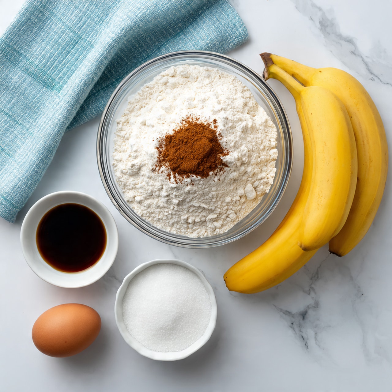 The image shows several baking ingredients neatly arranged on a white marbled surface. There is a large clear glass bowl filled with white flour and a small pile of brown cinnamon on top, positioned near the top center. To the bottom left, there is a single brown egg next to a small white bowl containing dark brown liquid, likely vanilla extract. Below these, there is a white bowl filled with granulated white sugar. Two ripe yellow bananas lie to the right side of the arrangement. A light blue striped kitchen towel is partially visible near the top left corner. Photo taken with an iphone --ar 4:5 --v 7
