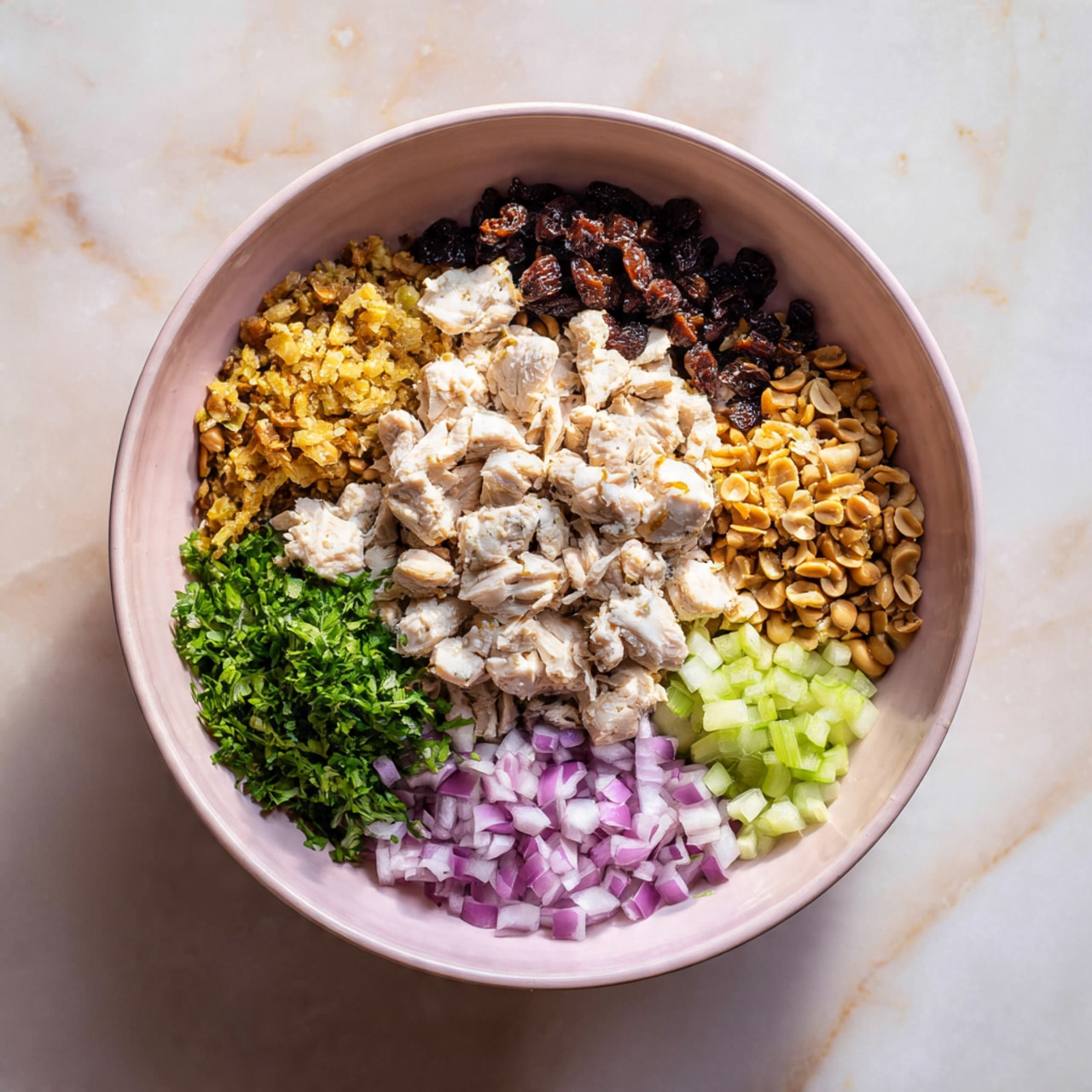 A large white bowl sits on a white marbled surface, filled with six separate layers of ingredients arranged in sections. The center holds chopped white chicken pieces, surrounded by crushed light brown peanuts on the right. Above the chicken are dark brown raisins, and below is a small bunch of fresh green chopped herbs. At the bottom left corner of the bowl are finely diced purple onions, while the bottom right corner contains small, pale green celery pieces. The bowl has a smooth texture and a soft pink color inside, contrasting with the fresh, colorful ingredients. Photo taken with an iphone --ar 4:5 --v 7