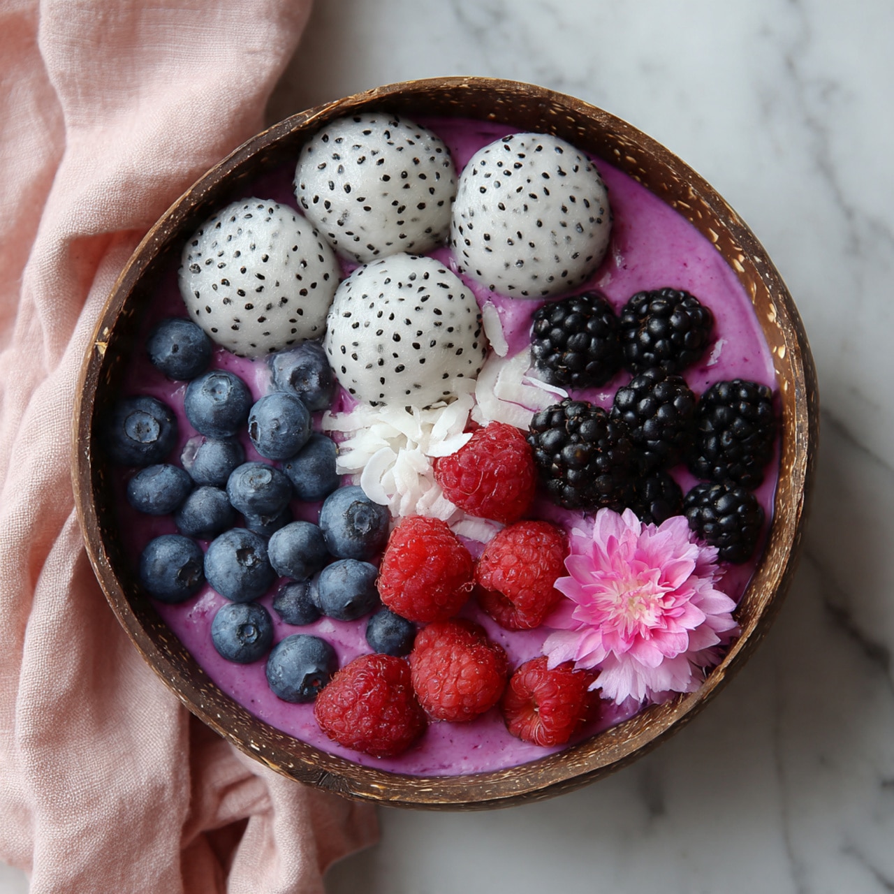 The image shows a rustic coconut shell bowl placed on a white marbled surface with a pink cloth underneath. Inside the bowl, the bottom layer is a smooth purple smoothie. On top of this layer, there are three different sections of fruit: on one side, round white dragon fruit balls with black seeds, next to them fresh blue blueberries stacked slightly, and on the other side, a mix of red raspberries and dark blackberries. There is also some shredded white coconut sprinkled over the fruit and smoothie parts. A small pink flower rests near the raspberries adding a delicate touch. The overall look is fresh, colorful, and natural. photo taken with an iphone --ar 4:5 --v 7