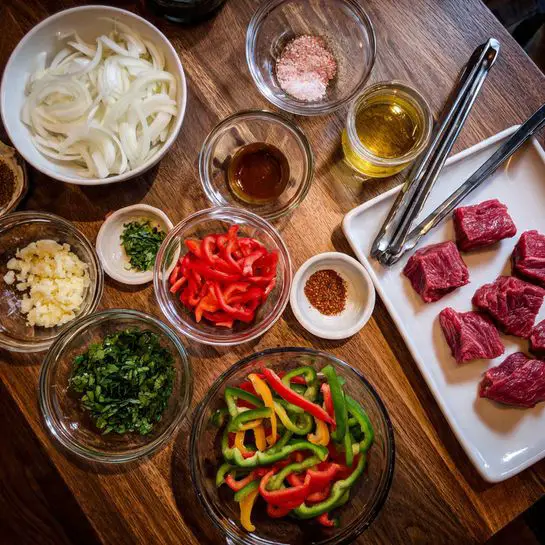 The image shows several clear glass bowls and white bowls arranged on a wooden table. One white bowl is filled with thinly sliced white onions, and another white bowl contains sliced red and green bell peppers with small chopped green herbs on top. Multiple small clear glass bowls hold minced garlic, chopped fresh herbs, pink salt, ground spices, tomato paste, olive oil, and a light brown liquid. To the right, a white plate holds several pieces of raw red meat, with metal tongs resting on the plate's edge. The scene is neatly arranged and ready for cooking preparation. Photo taken with an iphone --ar 4:5 --v 7
