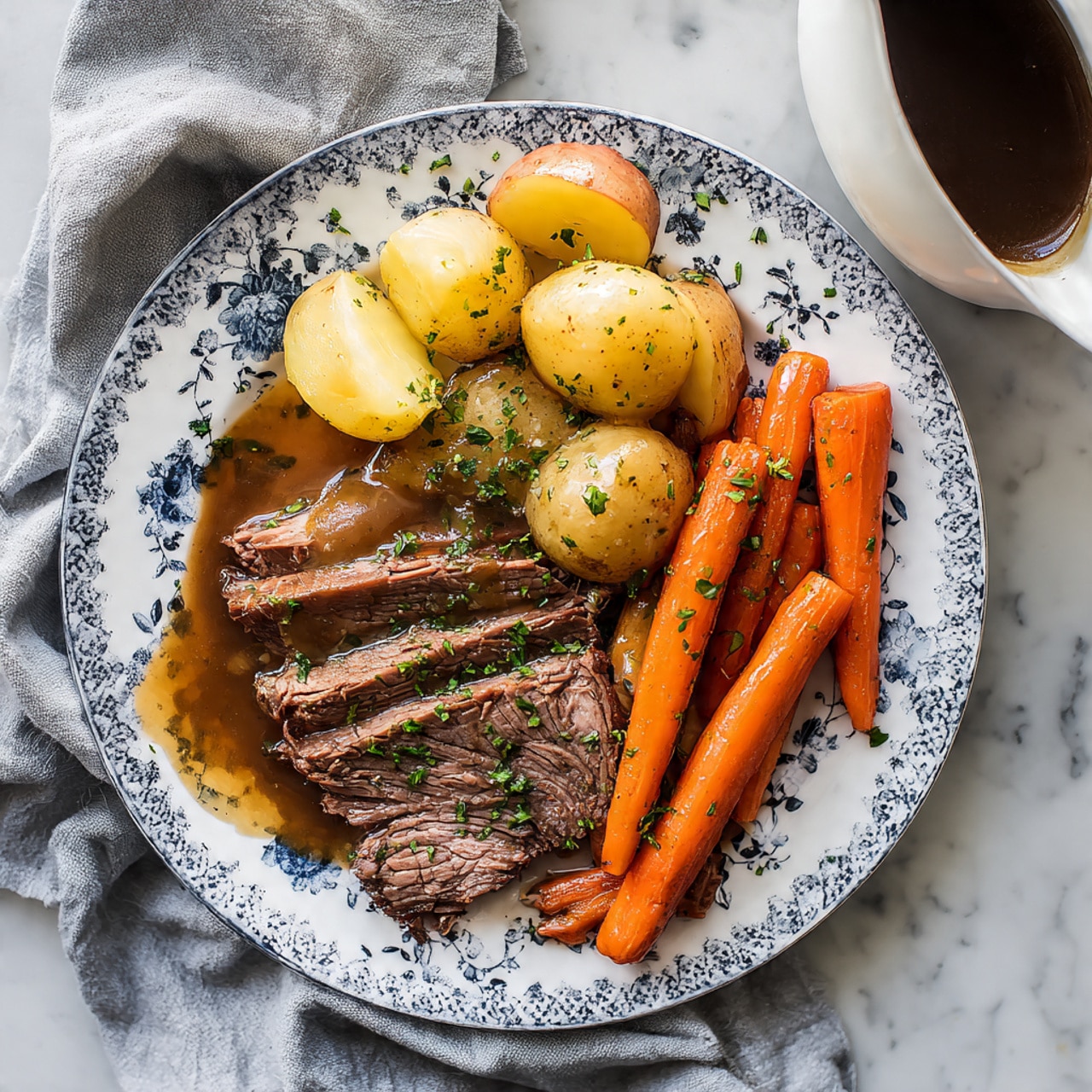 A white plate with blue floral patterns holds a serving of sliced brown roast beef layered in the center, surrounded by thick orange baby carrots and yellow chunks of potatoes. The beef looks juicy with a glossy brown sauce drizzled over it and sprinkled with chopped green herbs. The plate is placed on a white marbled surface with a soft gray cloth beside it. A small white gravy boat with dark sauce is partially visible on the right side. photo taken with an iphone --ar 4:5 --v 7