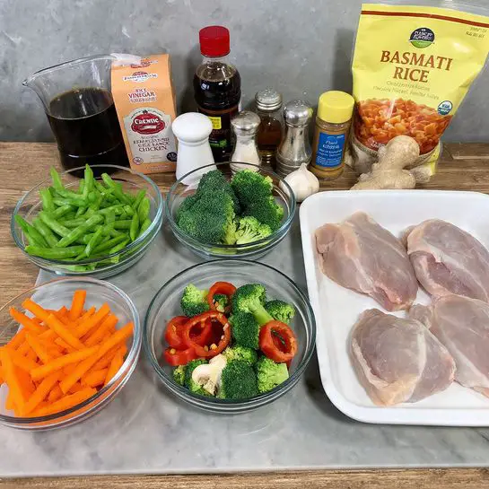 The image shows a wooden table with various cooking ingredients neatly arranged. At the front and center, there are clear glass bowls filled with bright green snow peas, vibrant orange carrot strips, fresh green broccoli florets, thinly sliced red bell pepper rings, and white mushrooms chopped into pieces. To the right, a white tray holds raw, pale pink chicken thighs labeled