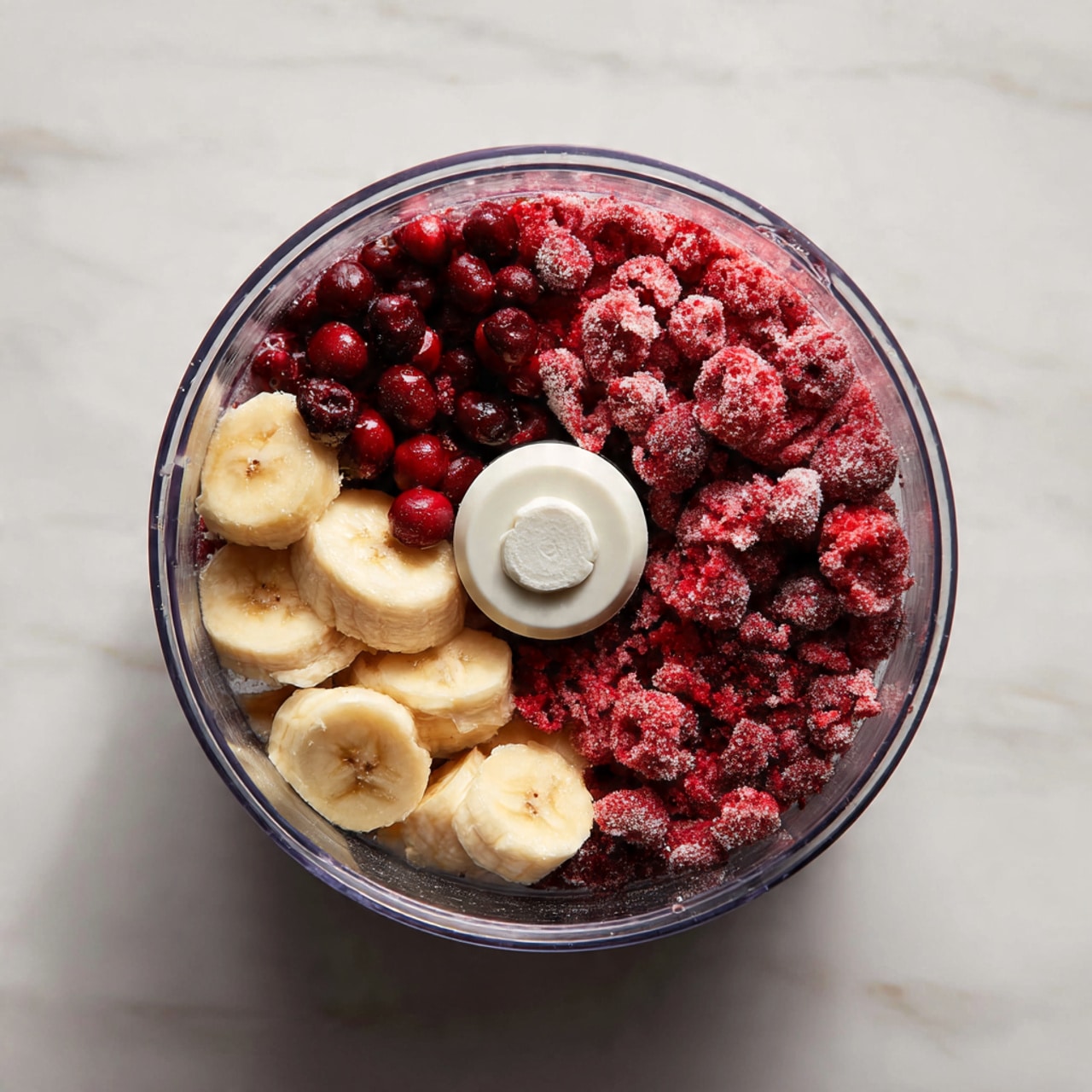 Inside a clear food processor bowl, there are two main layers of ingredients: on the right side, a bunch of frozen red berries with a rough, icy texture, and on the left side, several pieces of pale yellow banana, smooth and soft in appearance. A white circular blade piece is visible in the middle, separating the two layers slightly. The bowl sits on a white marbled surface, and next to it, there is a clear glass bowl with a white liquid. Photo taken with an iphone --ar 4:5 --v 7