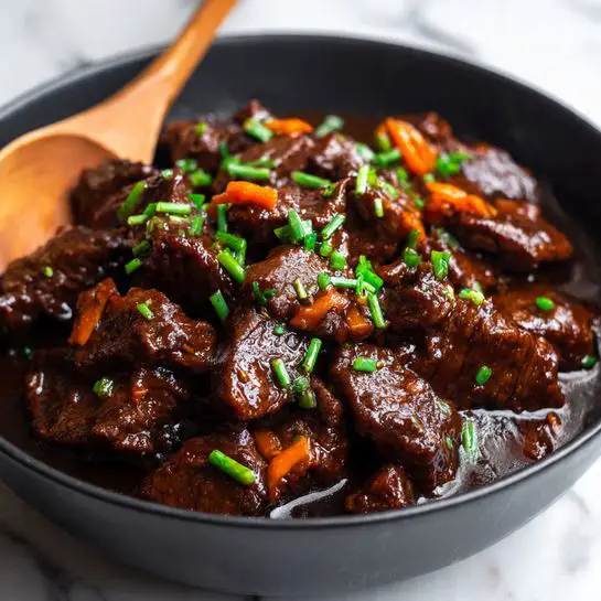 A close-up view of a dark black bowl filled with rich, shiny brown beef strips covered in thick sauce. There are small light green chopped scallions sprinkled evenly on top of the beef. Some orange carrot shreds are mixed in with the meat. The beef looks tender and glossy, sitting in the sauce at the bottom of the bowl. A wooden spoon is partly visible, resting inside the bowl beneath the beef. The background is a white marbled texture. Photo taken with an iphone --ar 4:5 --v 7