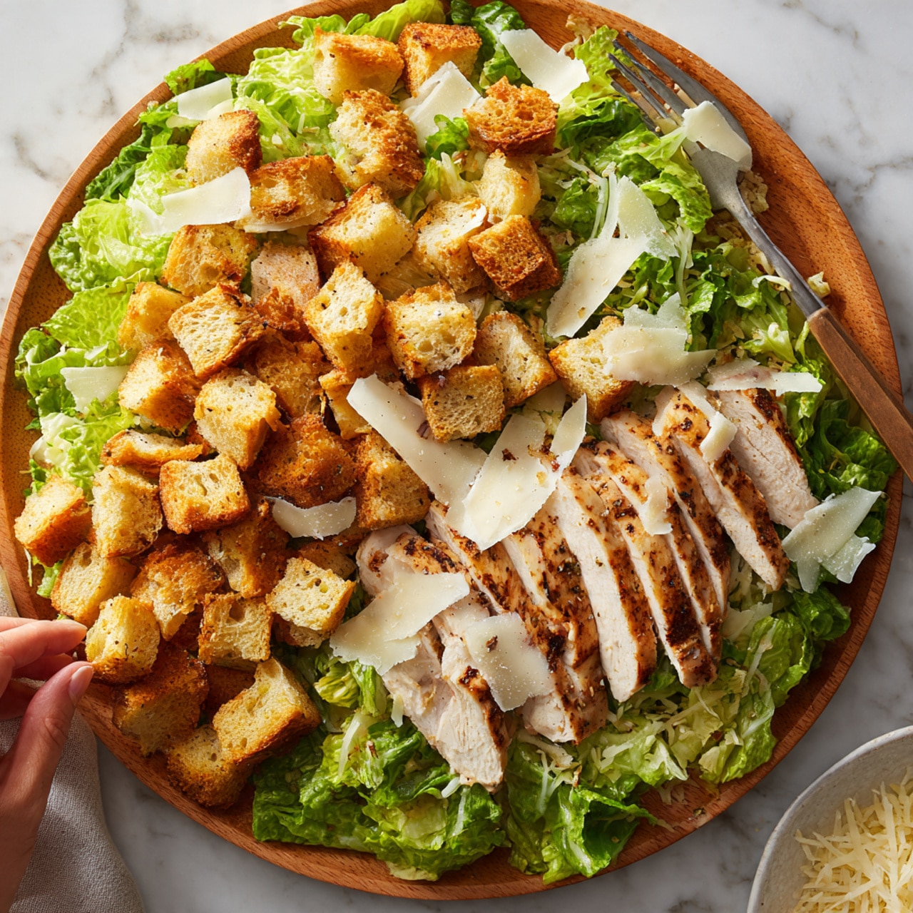A wooden plate holds a fresh salad with green lettuce leaves spread across the bottom. On top, there are many golden-brown toasted bread cubes scattered evenly. In the center, slices of grilled chicken with a light brown color and visible grill marks are placed neatly in one layer. Thin shreds of white cheese are sprinkled over the salad, adding a soft texture. A white marbled surface serves as the background, and a woman's hand is seen holding a fork near the plate. Photo taken with an iphone --ar 4:5 --v 7