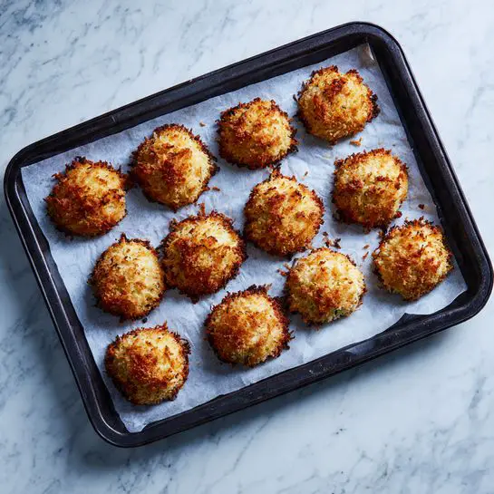 The image shows a dark baking tray with 11 pieces of baked food placed evenly on it. Each piece is coated with a golden-brown crust that looks crispy and crumbly on top. The tray is lined with white paper, and it rests on a white marbled surface. There is no other item or decoration in the picture. The overall look is clean and simple. photo taken with an iphone --ar 4:5 --v 7