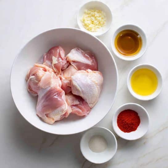 The image shows two raw chicken breasts placed side by side on crumpled white parchment paper in the center, lying on a white marbled surface. Around them are various ingredients: on the top left, a wooden pepper grinder, a clove of garlic, a small block of cheese, and a shallot. Near the chicken, there are small bowls with white salt and dried herbs on the upper right. Below the chicken, there is a glass mug filled with light yellow broth, a blue tube of anchovy paste, a small stone bowl filled with green capers, a glass of clear white wine, a metal measuring cup filled with flour, and two bright yellow lemons. Fresh green parsley leaves are at the lower left corner. At the top, a container of buttermilk and a pepper shaker complete the set. The photo taken with an iphone --ar 4:5 --v 7