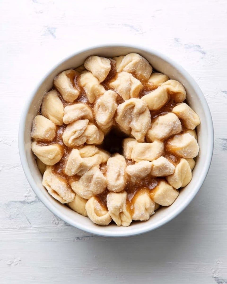 The image shows a white bundt pan filled with small pieces of dough that are light golden brown and have a soft texture. The dough pieces are arranged in a thick layer, filling the pan almost to the top, with some pieces showing slight folds and creases. The pan is placed on a white marbled surface with a subtle clean look around it. The scene gives a fresh baking feeling, and the light highlights the smooth and slightly shiny sides of the dough pieces, making them look fresh and ready for baking. Photo taken with an iphone --ar 4:5 --v 7