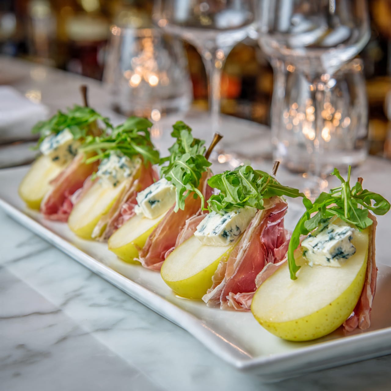 The image shows several pieces of food arranged on a dark slate board placed on a white marbled surface. Each piece is a small pear slice at the bottom, with a layer of soft white cheese on top, all wrapped with a thin slice of pink prosciutto secured by a wooden toothpick with a dark tip. The pear slices have smooth, light yellow skin with a touch of reddish blush on one side, while the cheese layer inside is creamy and soft. The prosciutto looks thin and slightly shiny, with natural folds, adding texture and color contrast to the arrangement. In the background, blurred whole pears with red and green skin are visible. A woman's hand is seen adjusting one of the bites. The lighting is soft and natural, highlighting the colors and textures. Photo taken with an iphone --ar 4:5 --v 7