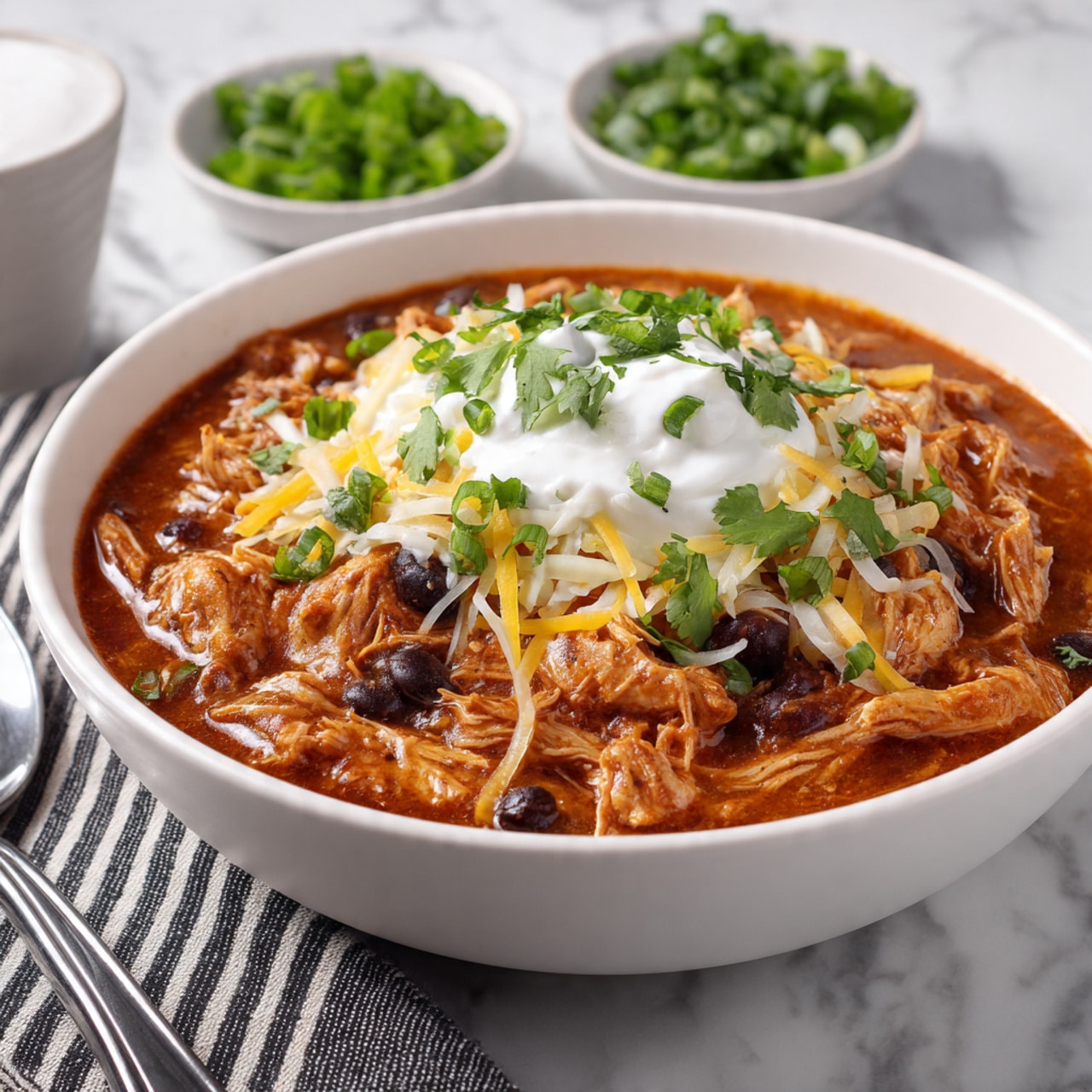 A deep white bowl filled with a thick, orange-brown stew with chunks of shredded chicken and black beans mixed throughout the rich sauce. On top, there is a layer of white sour cream surrounded by a layer of finely shredded yellow and white cheese sprinkled with chopped green onions and fresh chopped cilantro. The bowl sits on a white marbled surface with blurred small white dishes filled with extra chopped green onions and sour cream in the background, along with a striped black and white cloth napkin and a silver spoon to the left. photo taken with an iphone --ar 4:5 --v 7