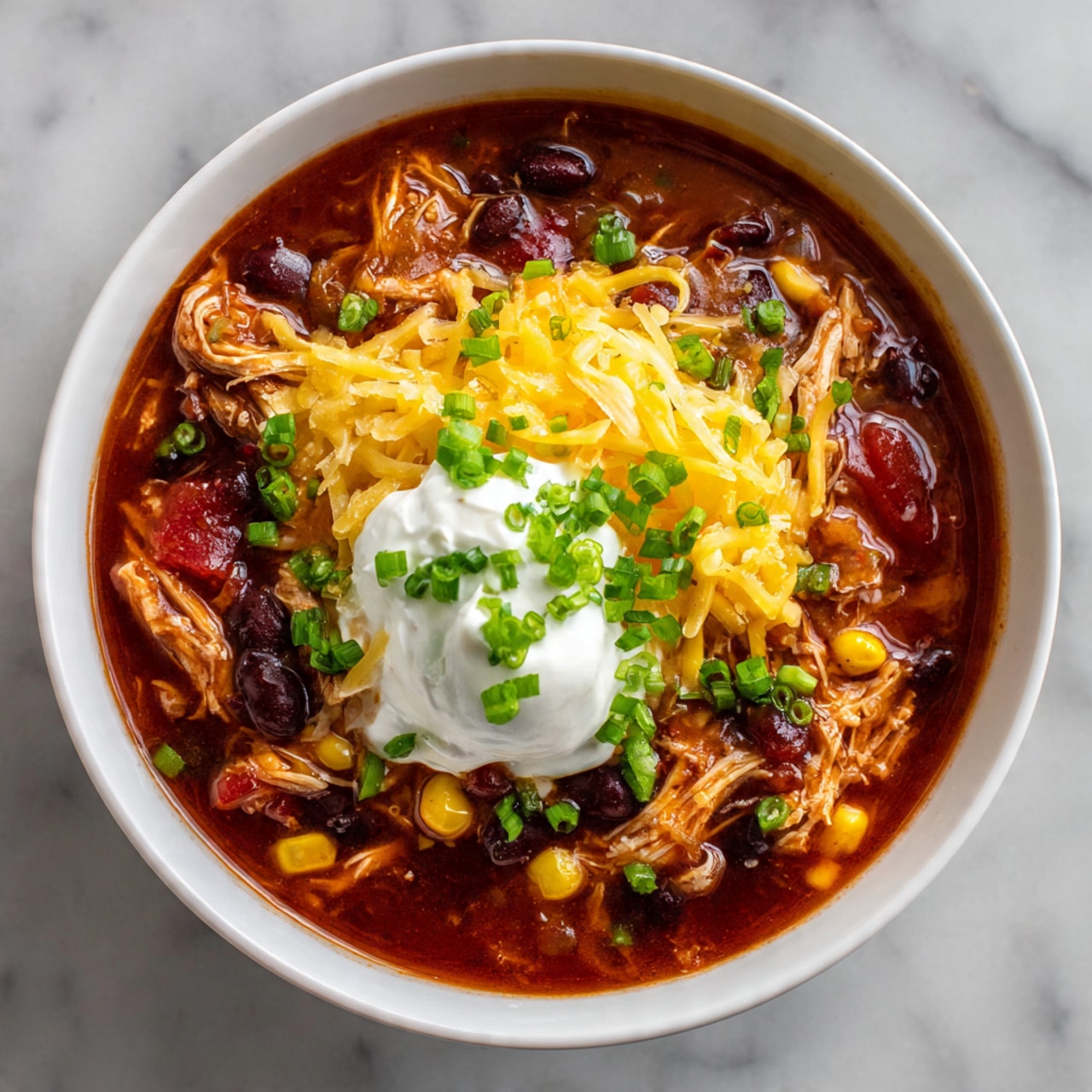 A white bowl filled with a thick stew made of shredded chicken, black beans, kidney beans, corn, and diced tomatoes in a reddish broth, topped with a dollop of white sour cream, a pile of shredded yellow cheddar cheese, and sprinkled with small green chopped scallions. The bowl sits on a white marbled surface. photo taken with an iphone --ar 4:5 --v 7