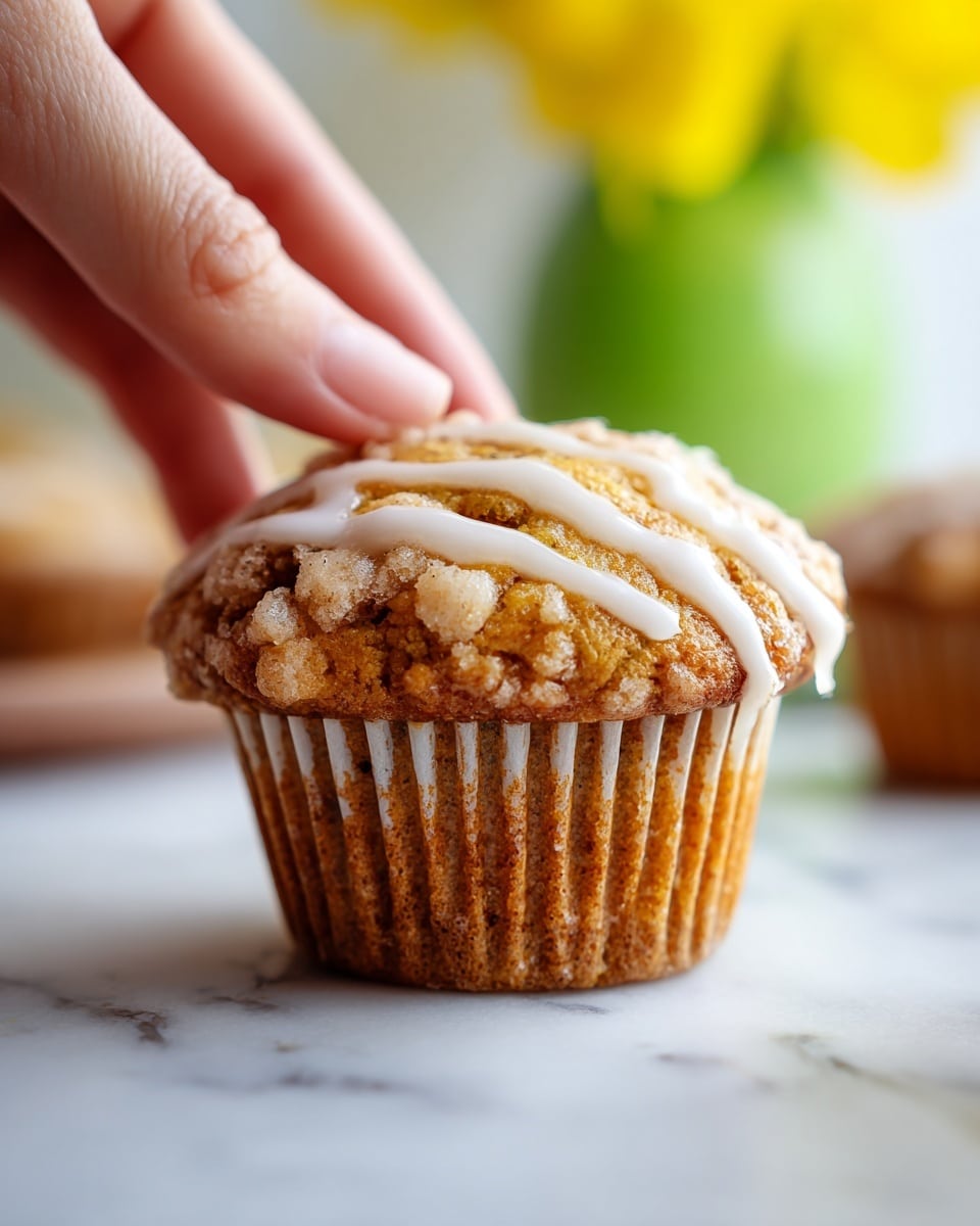 The image shows a single muffin placed on a white marbled surface. The muffin has a light brown, crumbly top with a drizzle of white icing that runs down slightly over the edges. The muffin liner is slightly wrinkled and white. The background is bright and softly blurred with a green and yellow shape in the distance. A woman's hand is gently holding the muffin from the side. Photo taken with an iphone --ar 4:5 --v 7