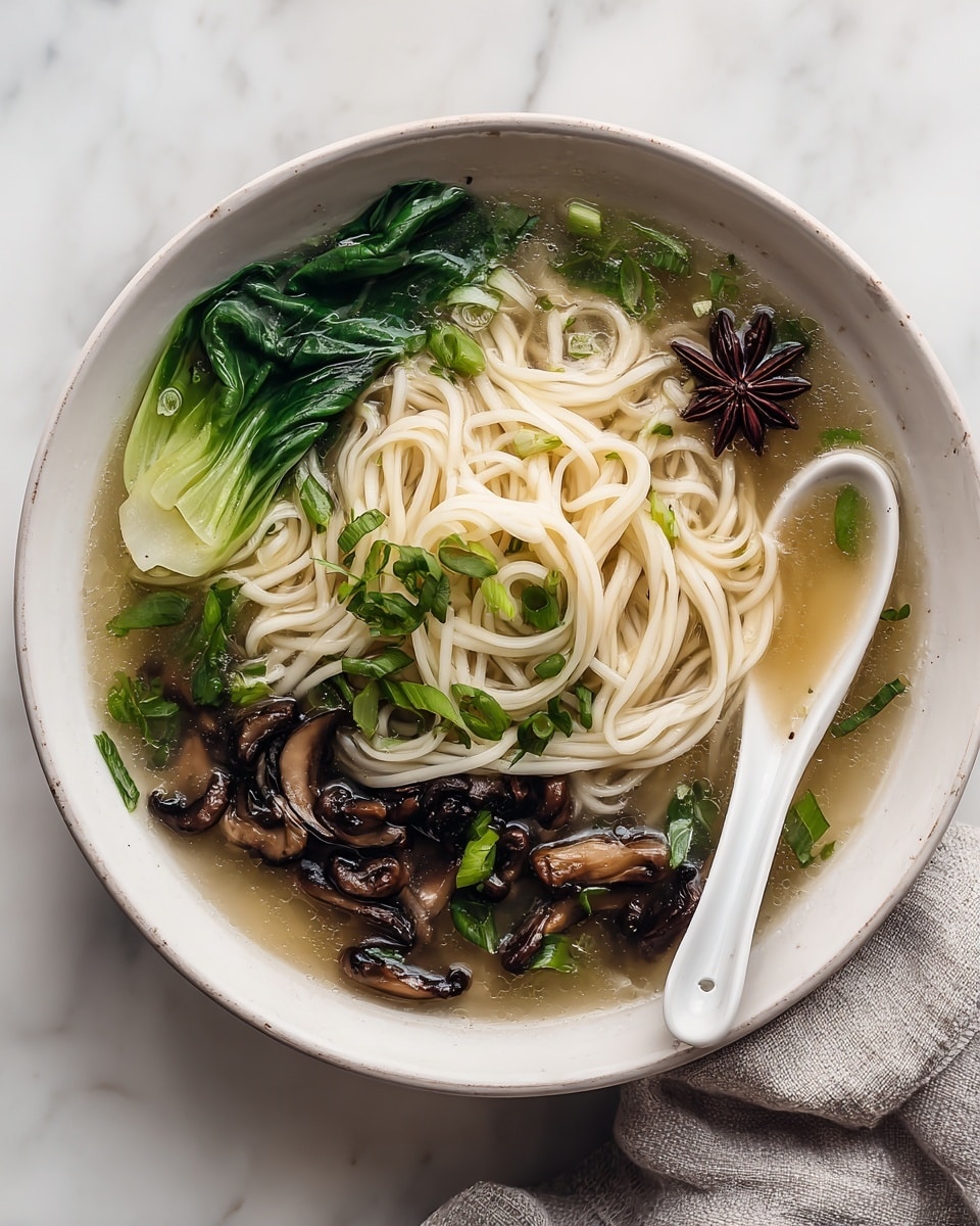 A white bowl filled with a clear broth containing thick, white noodles arranged loosely in the center. Around the noodles are dark brown, sliced mushrooms with a soft texture, bright green leafy vegetables, and chopped green onions floating on the surface. A dark brown star anise piece is visible near the noodles. A white soup spoon is placed inside the bowl on the right side, resting in the broth. The bowl is on a white marbled surface with a light gray cloth partially visible on the right side. photo taken with an iphone --ar 4:5 --v 7