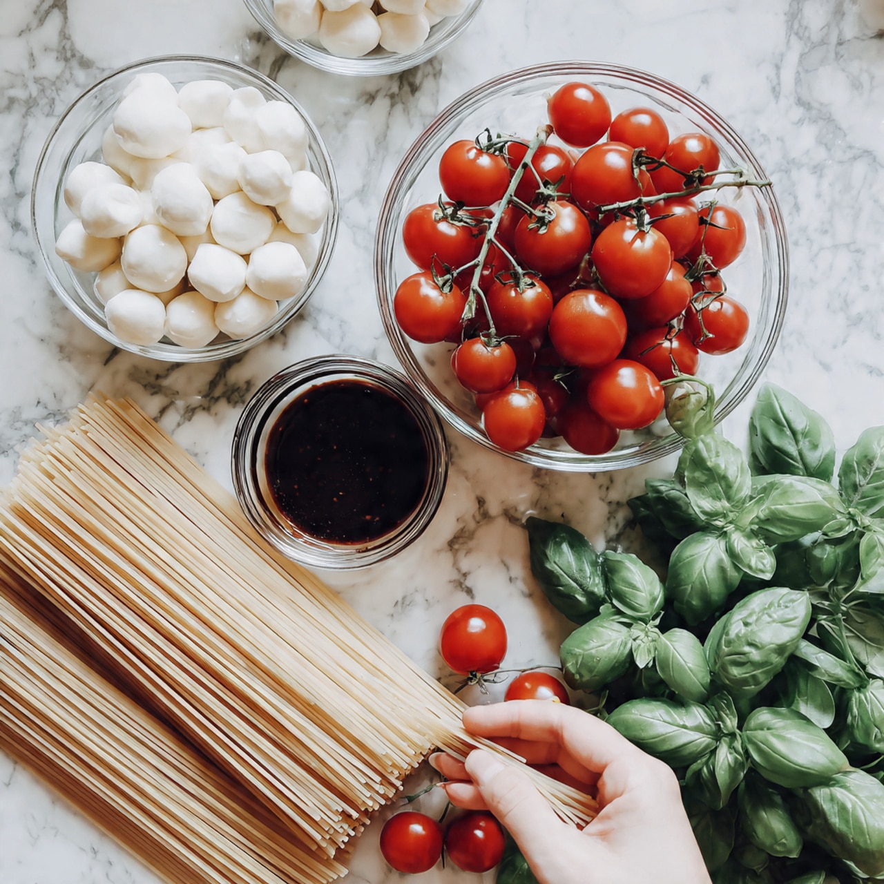 The image shows four clear glass bowls arranged on a white marbled surface. The largest bowl is filled with bright red cherry tomatoes still on the vine, sitting at the top right. Below and slightly to the left, there is a medium bowl holding fresh green basil leaves with visible texture on the leaves. To the left, a smaller bowl contains smooth white mozzarella balls. At the top left, there is a small bowl with dark balsamic vinegar. Next to the bowls on the left side, there is a small stack of uncooked pale, light beige fettuccine pasta sticks tied gently. The lighting is soft and natural, making the colors of the ingredients look fresh and vibrant, photo taken with an iphone --ar 4:5 --v 7