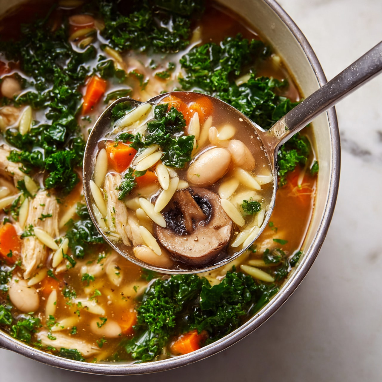 The image shows a close-up of a clear soup filled with many ingredients. It has visible layers including light brown broth as the base, scattered bright green leafy kale, small chunks of orange carrot, slices of light brown mushrooms with darker edges, white beans, and short white noodles resembling orzo. The soup has pieces of light beige chicken mixed in, and the textures look soft and cooked. A shiny silver ladle is scooping some of the soup, showing the mix of vegetables and broth clearly. The background is a white marbled surface. photo taken with an iphone --ar 4:5 --v 7