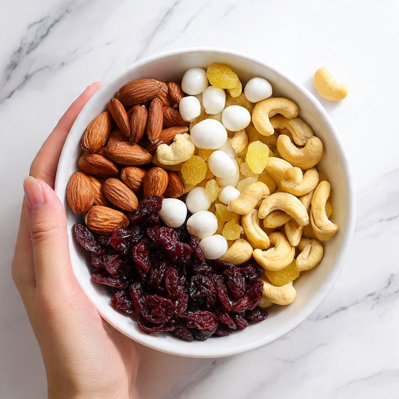 A white bowl filled with a mix of dry fruits and nuts arranged closely together. The bottom layer is made of dark red dried cranberries, giving a rough and wrinkled texture. On top, there are smooth, light brown almonds and large, curved yellowish cashews spread evenly. Scattered among the nuts and berries are round, white yogurt-covered raisins, adding a soft shiny texture. The bowl is set on a white marbled surface, with a woman's hand visible holding the bowl from the top edge. photo taken with an iphone --ar 4:5 --v 7