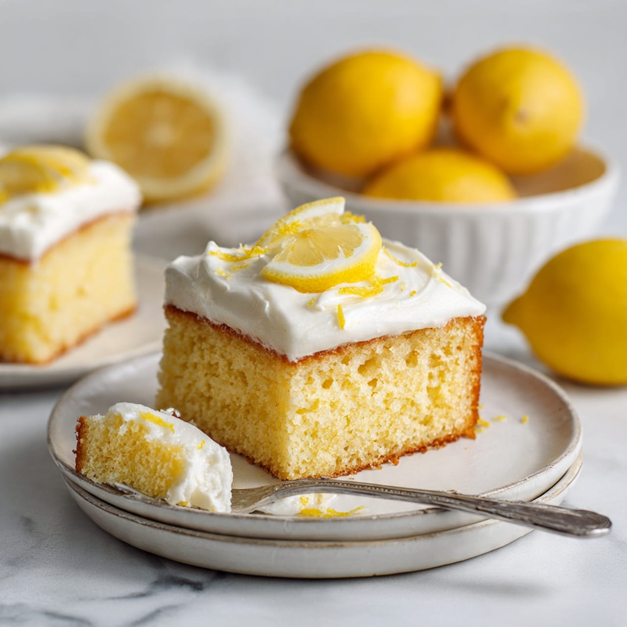 A square piece of lemon cake sits on a stack of two white plates with a white marbled surface beneath. The cake has two clear layers: a moist, yellow sponge base and a thick white cream layer topped with a small lemon slice and some lemon zest. A silver fork holds a bite of the cake with cream on it, placed on the plate near the cake piece. In the background, there is another white plate holding a similar piece of cake and a white bowl with three whole bright yellow lemons. The scene is bright and clean, showing a fresh and simple dessert. photo taken with an iphone --ar 4:5 --v 7