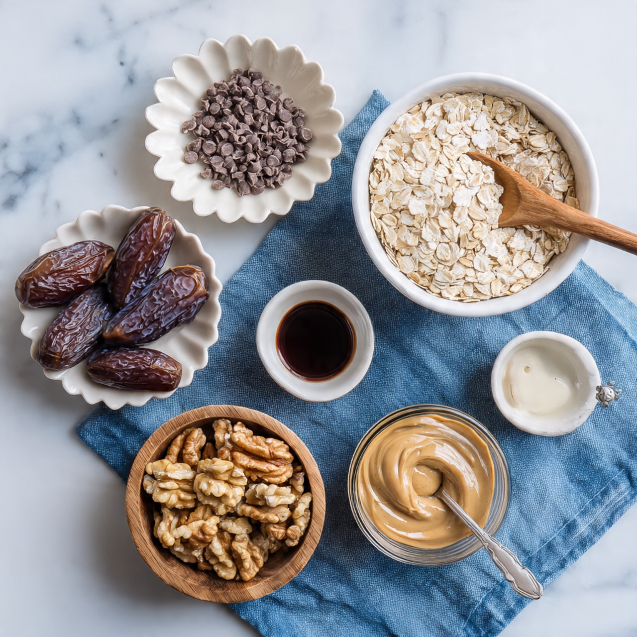 The image shows six bowls of ingredients arranged on a white marbled surface with a blue cloth nearby. The top right bowl is white and filled with light beige rolled oats with a wooden spoon resting inside. Below it to the right is a small glass bowl of smooth brown almond butter with a spoon in it. To the bottom right of that is a tiny white bowl containing dark brown vanilla extract. Below on the bottom left side is a light wooden bowl filled with light brown walnut halves. Above that is a speckled white bowl holding shiny dark brown whole dates. At the top left is a white bowl with scalloped edges filled with small dark brown chocolate chips. Photo taken with an iphone --ar 4:5 --v 7