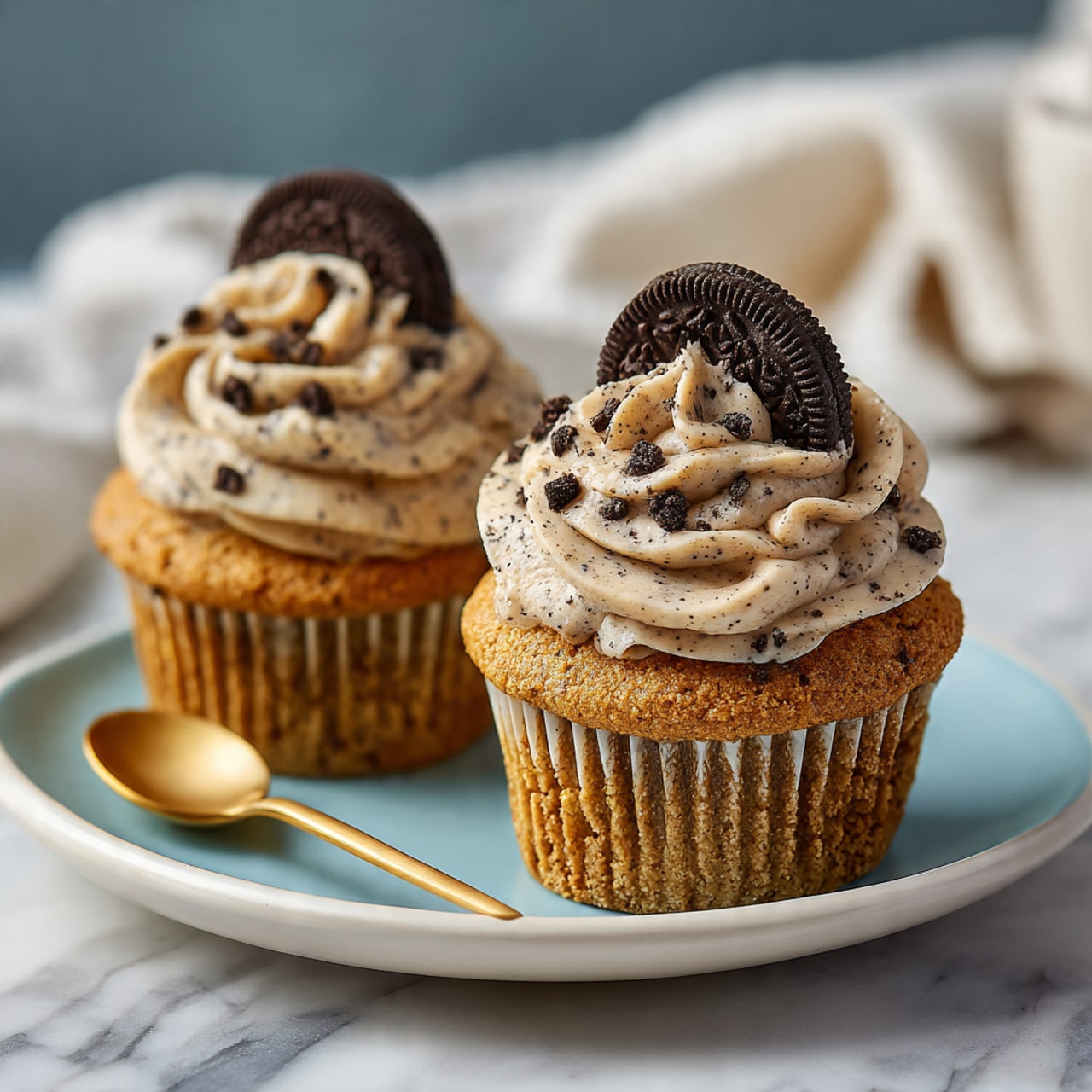 The image shows two cupcakes on a white plate with a light blue center. Each cupcake has a golden brown base with a soft texture. On top, there is a thick layer of creamy, light beige frosting with small dark specks, swirled in a circular pattern, creating a fluffy and textured look. Each cupcake is decorated with half of a small chocolate sandwich cookie placed upright in the center of the frosting. The background is a white marbled surface with a blurred gold spoon and white cloth in the back. Photo taken with an iphone --ar 4:5 --v 7