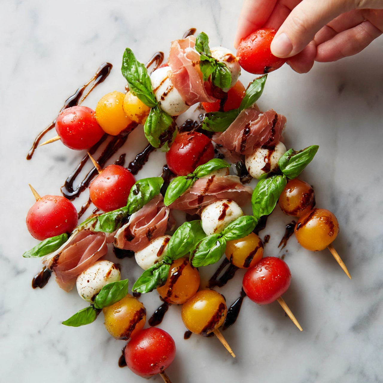 The image shows a white plate filled with small appetizers on wooden skewers arranged in a casual pile. Each skewer has four layers: a bright green basil leaf at the bottom, a shiny red or yellow cherry tomato on top of it, followed by a white mozzarella ball with a smooth texture, and topped with thin slices of pink cured meat. The skewers are drizzled with dark balsamic glaze in a zigzag pattern. The plate is placed on a white marbled surface. A woman's hand is reaching from the left side to pick one. Photo taken with an iphone --ar 4:5 --v 7