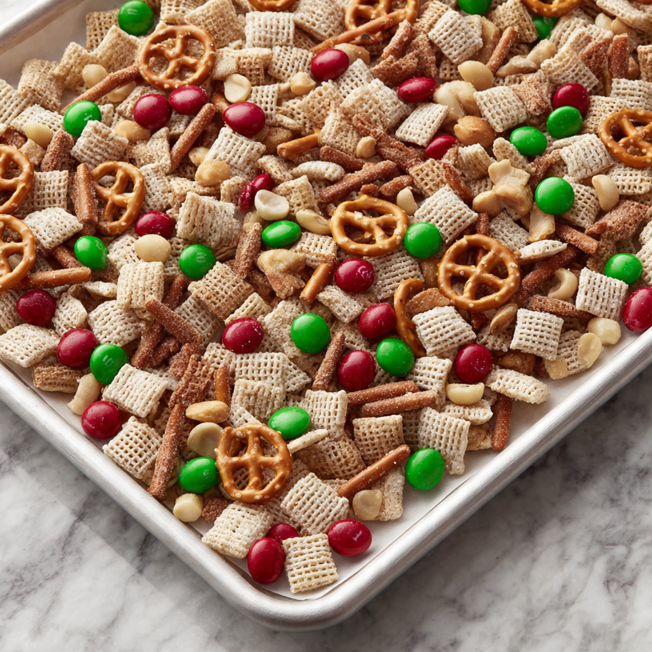 A white tray lined with parchment paper is filled with a colorful snack mix spread in one even layer. The mix has light beige square cereal pieces with a grid texture, small round cereal loops in a similar beige shade, and crunchy pretzel sticks in a deeper brown color. Scattered throughout are bright red and green candy-coated chocolates, and light brown peanuts. The whole mix is lightly coated in a white, sugary layer that gives some pieces a frosted look. The background shows a white marbled surface. Photo taken with an iphone --ar 4:5 --v 7